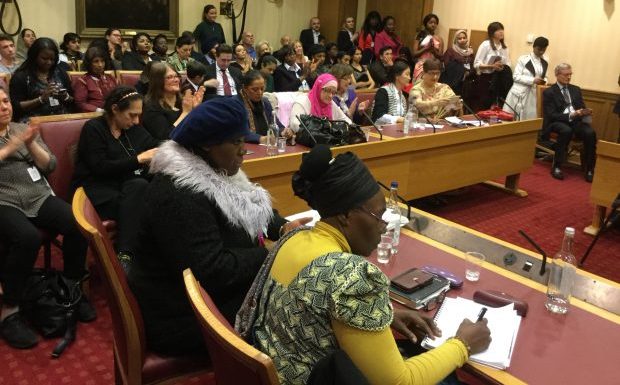 A multi-ethnic group of women are sitting at desks in a room, listening to a speaker.