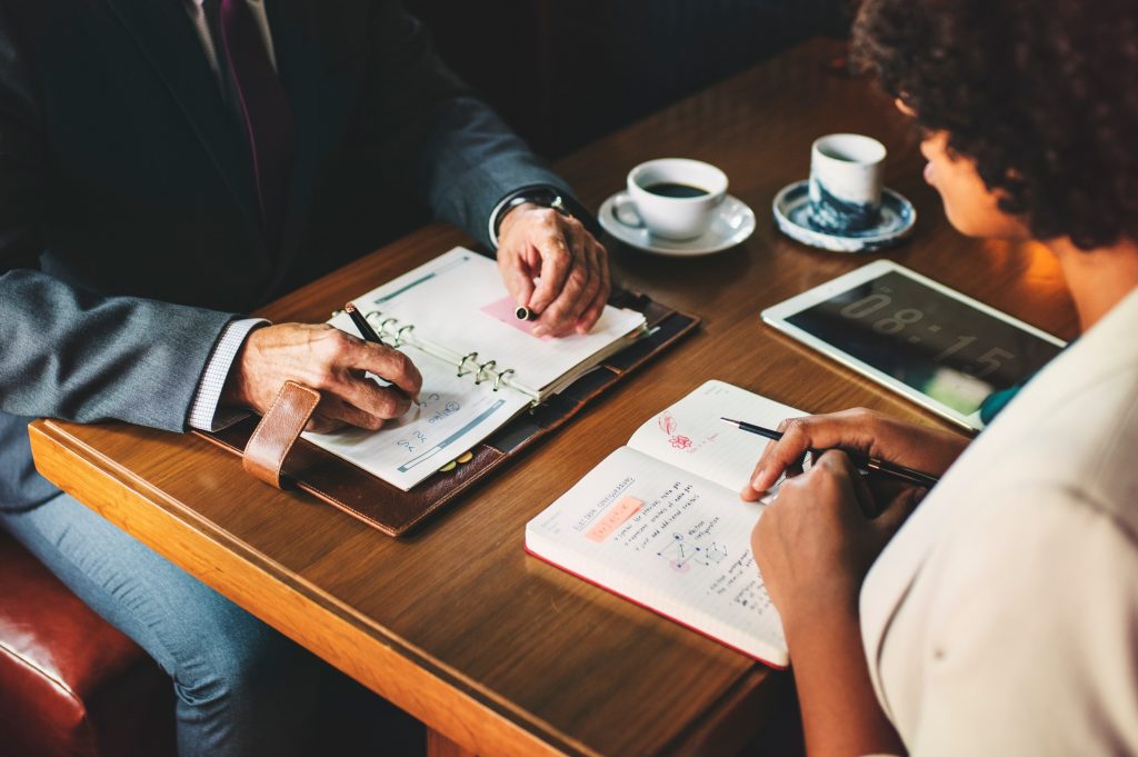 A man in a suit and a woman sat opposite each other at a table with diaries and note books