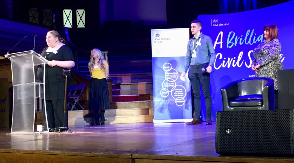 4 people standing on the stage with one speaking at a podium in front of a banner saying "A Brilliant Civil Service"