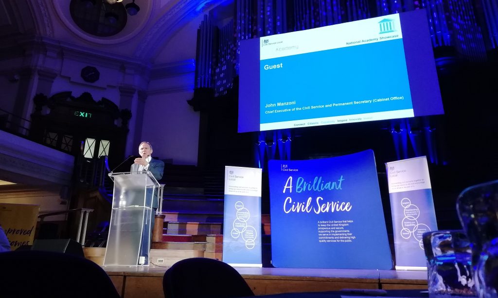 John Manzoni speaking at a podium on stage at the showcase event in front of a large banner which reads "A Brilliant Civil Service"