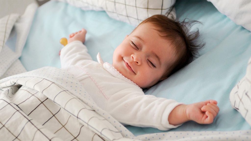 Baby sleeping on its back with arms out to its side and smiling with a checked blanket mid way up its body