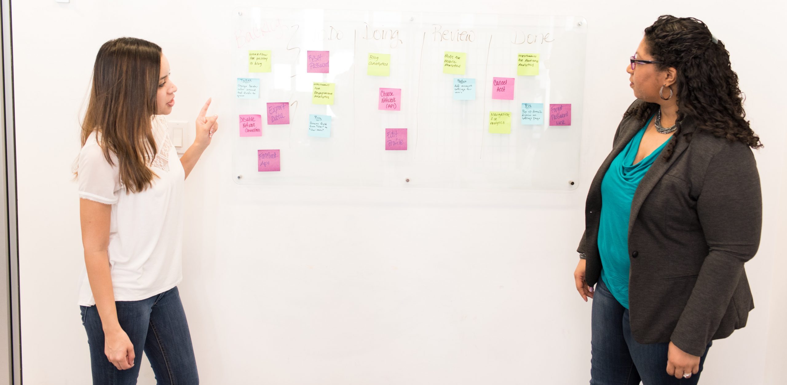 Two women standing by a white board with post-it notes saying various words relating to continuous improvement