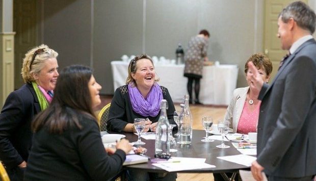 Three women are sat around a table talking and laughing. Aman is stnding next to then with his back to the camera.