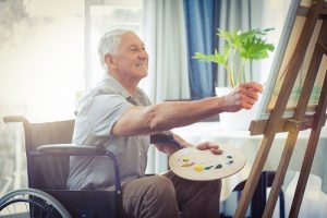 elderly man sitting in a wheelchair holding a paint brush in his left hand and leaning towards an easel with a paint pallet in his left hand. , the elderly man is reaching out towards an easel painting. 